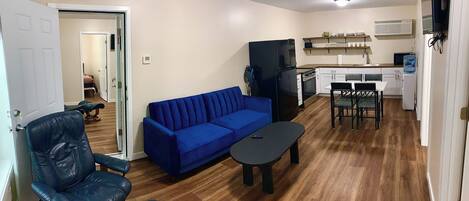 Un espacio habitable compacto con un sofá azul, un sillón de cuero negro, una pequeña mesa de comedor y una zona de kitchenette.