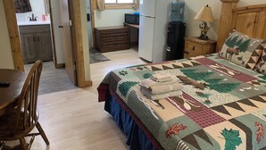 Ein gemütliches Zimmer mit einem Bett, einer Holzkommode, einem Kühlschrank, einem Wasserspender und einer Essecke mit Tisch und Stühlen.