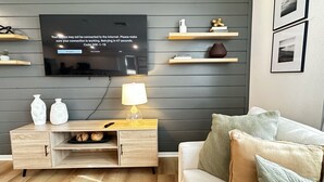 Una sala moderna con un televisor de pantalla plana montado en la pared, una mesa consola de madera con una lámpara y un sofá con cojines.