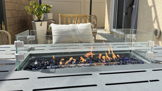 Una chimenea de vidrio con piedras y llamas, rodeada por un patio con una silla, una mesa y una planta en maceta.
