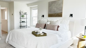 Un dormitorio con una cama grande, una mesita de noche con lámpara, una estantería con objetos decorativos y una ventana.