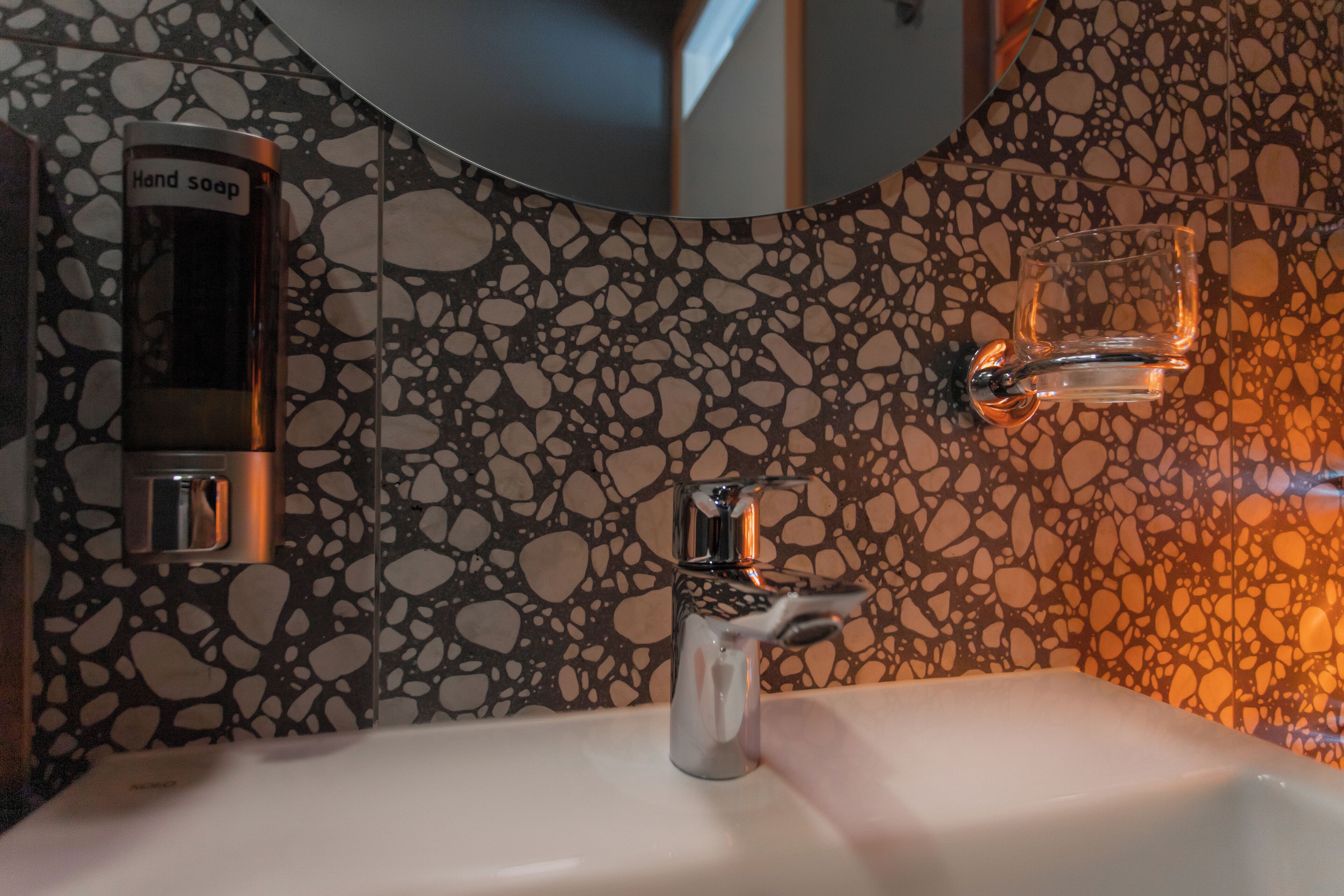A bathroom with a sink, a hand soap dispenser, and a glass soap dish on the wall.