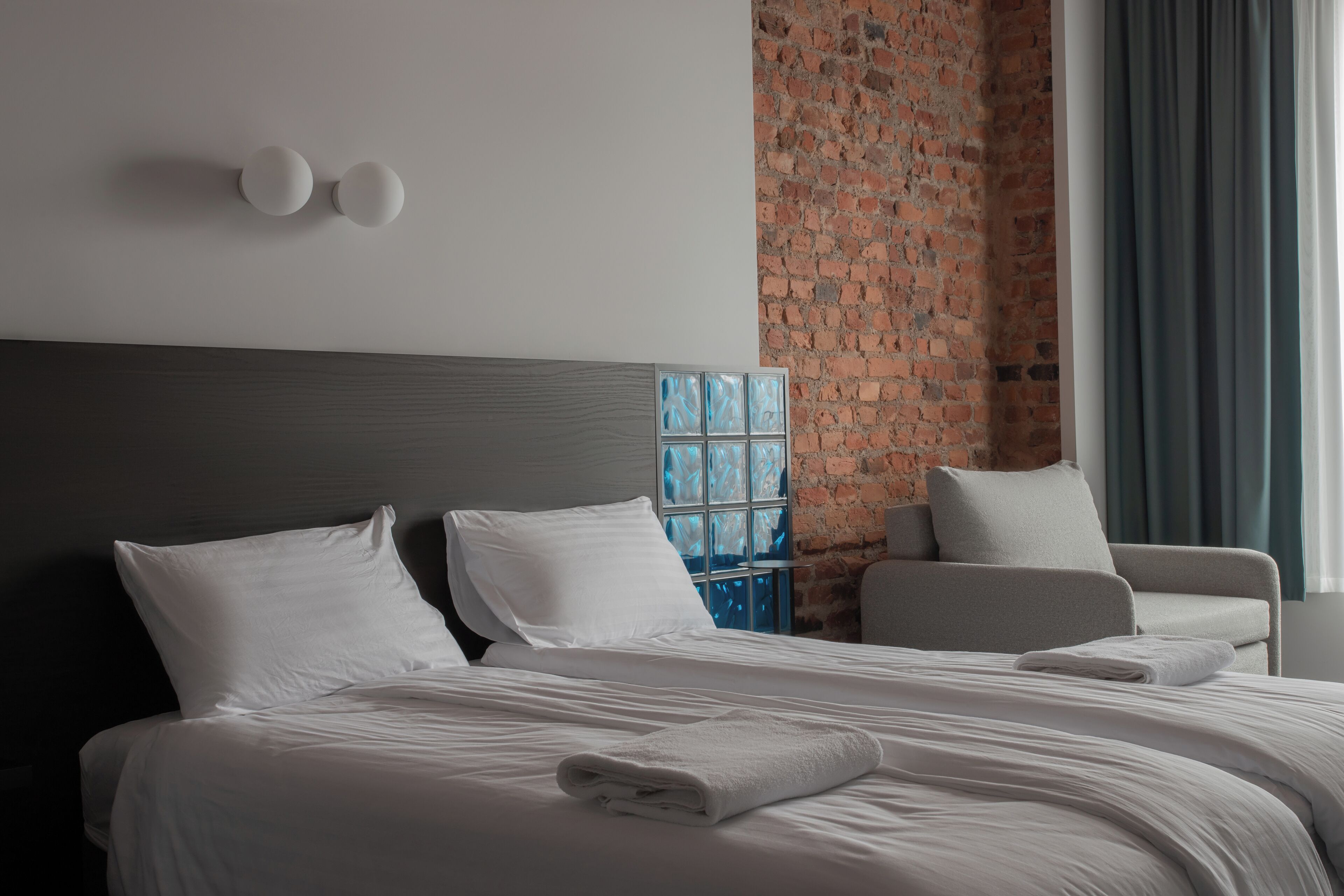 Een moderne hotelkamer met een groot bed, een grijze fauteuil en een bakstenen muur.