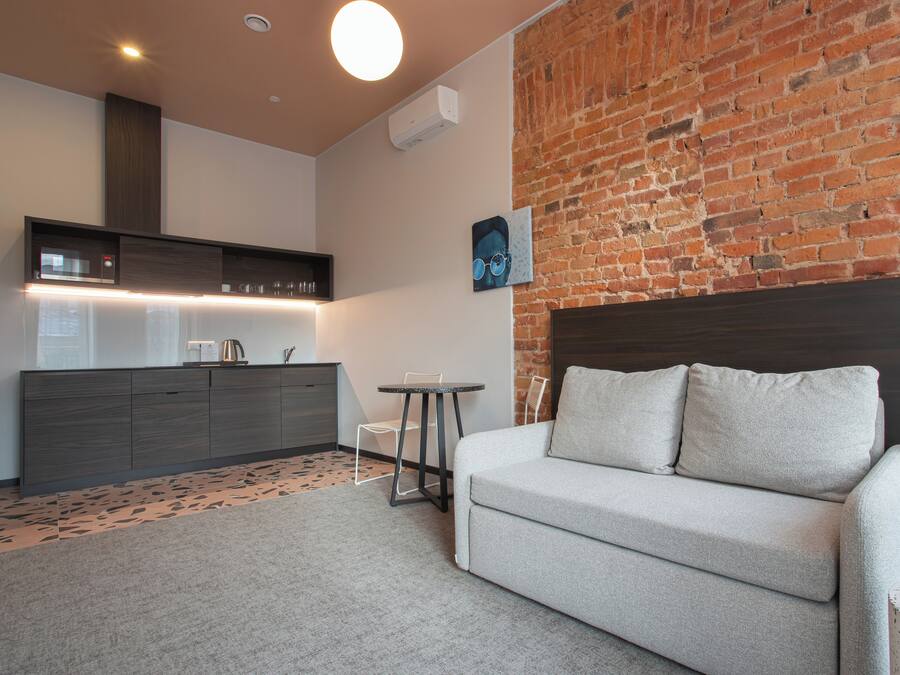 A modern living room with a grey sofa, a small round table, and a kitchen area with a built-in oven and a brick wall.