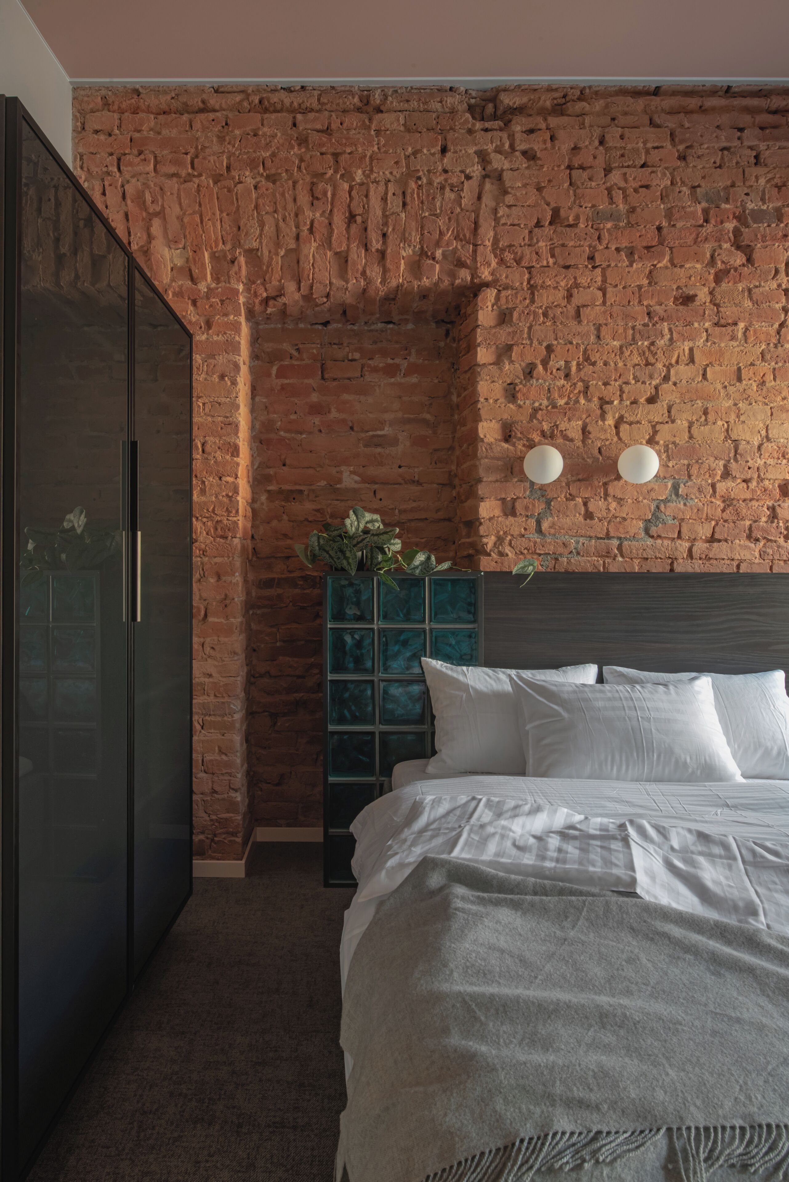 A bedroom with a brick wall, a dark wardrobe, a bed with white bedding, and a green cabinet with plants.