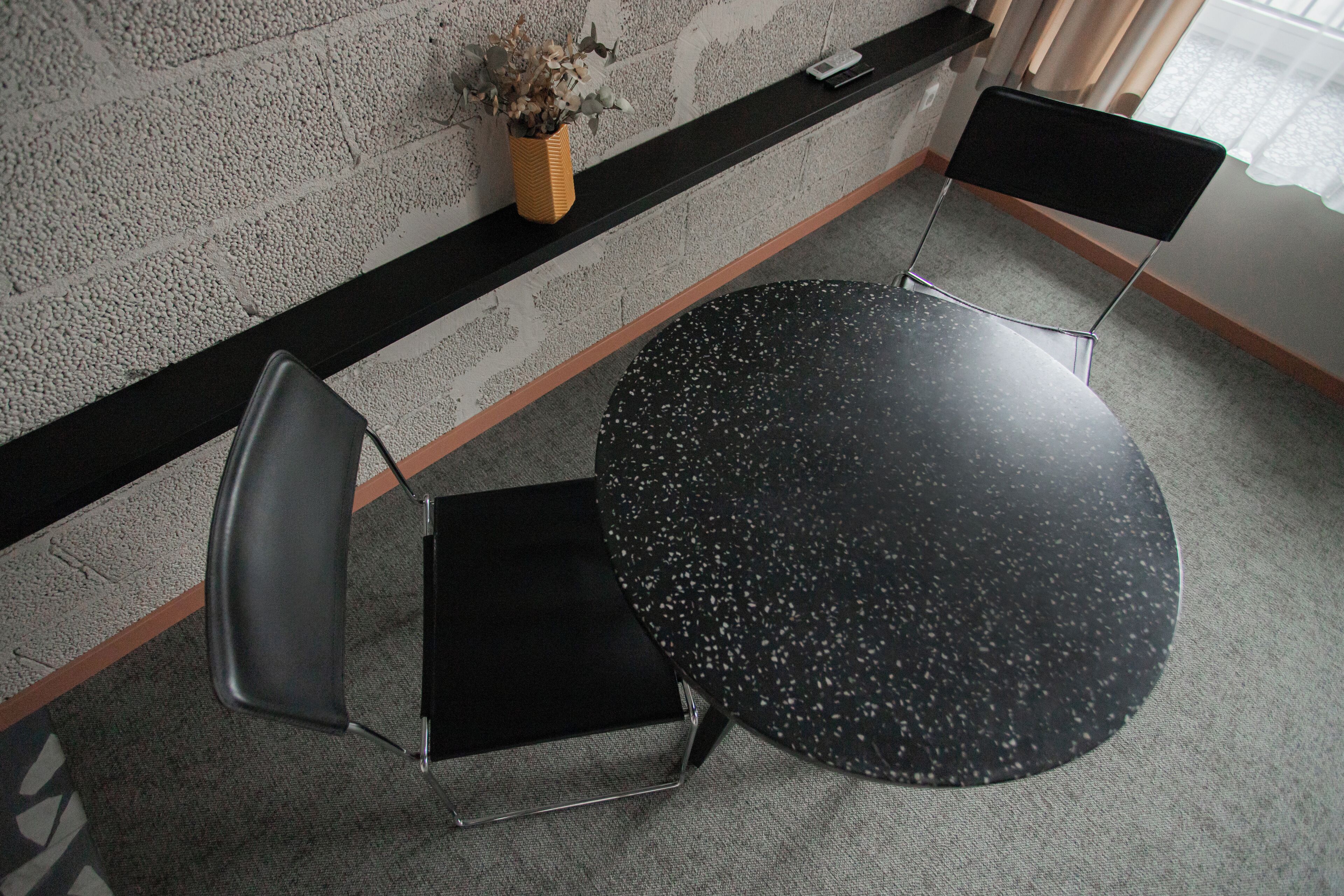 A small, round, black table with a speckled surface, a black chair with a metal frame, and a vase with dried flowers on a black shelf against a textured wall.