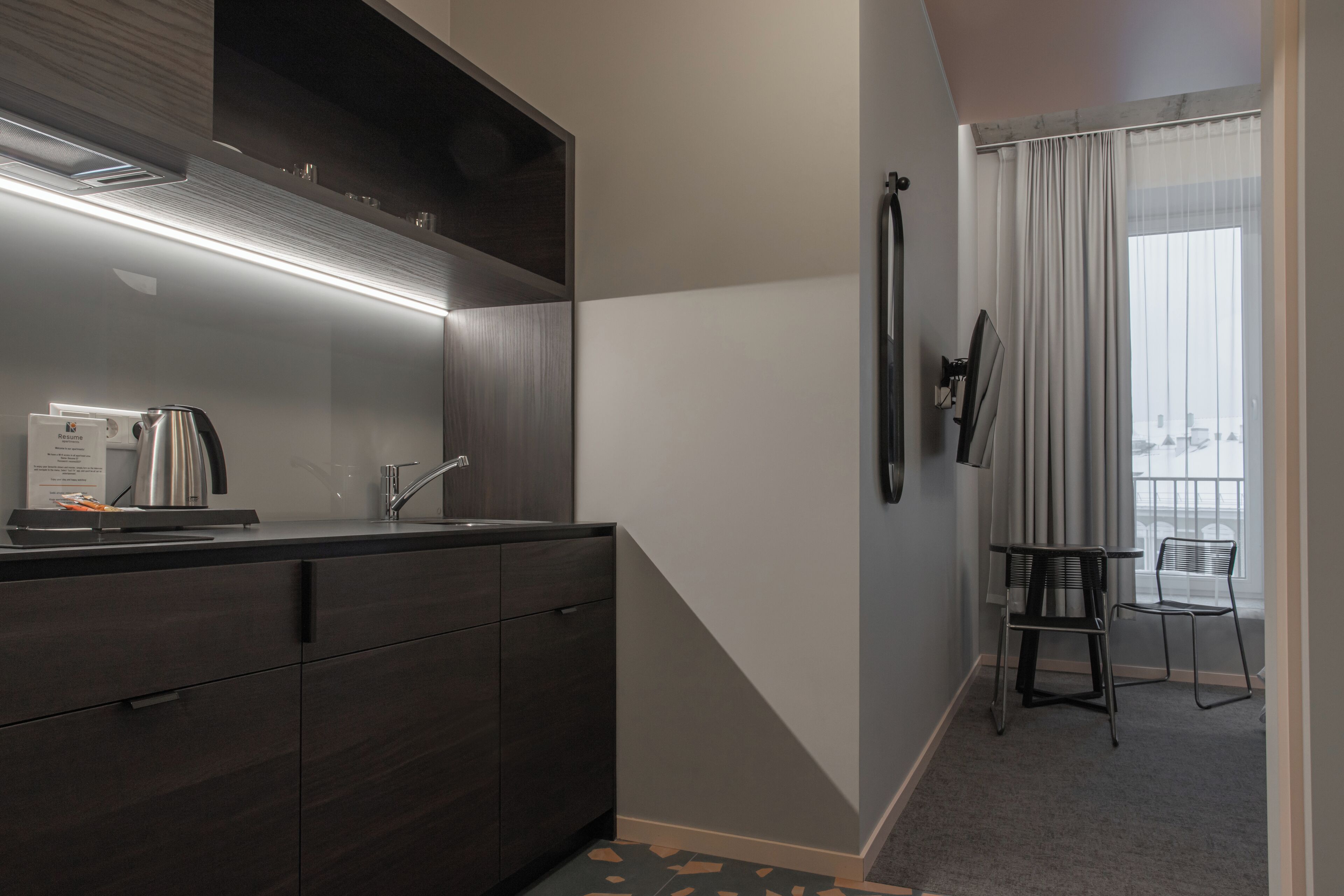 A modern kitchen with dark cabinetry, a stainless steel kettle, and a built-in oven. There is a small seating area with a chair and a table near a large window with sheer curtains.