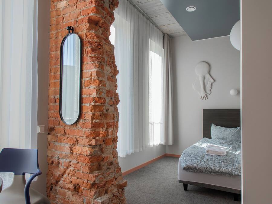 A modern hotel room with a brick accent wall, a bed with a grey headboard, a large mirror, and a view of the outdoors.