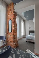A modern hotel room with a brick accent wall, a bed with a grey headboard, a large mirror, and a view of the outdoors.