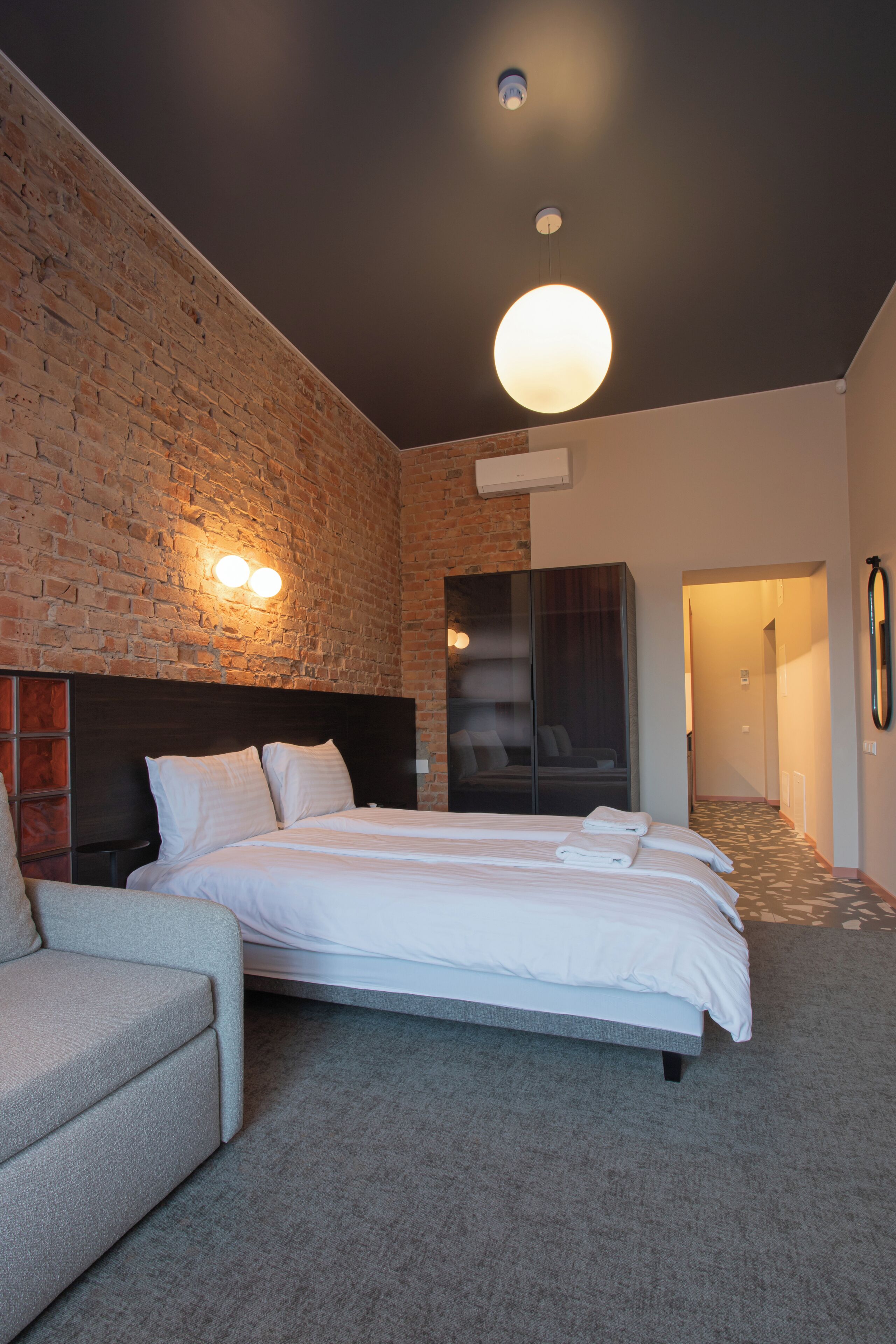 A modern hotel room with a brick accent wall, a large bed with neatly made linens, a seating area with a sofa, and a hallway leading to another room.