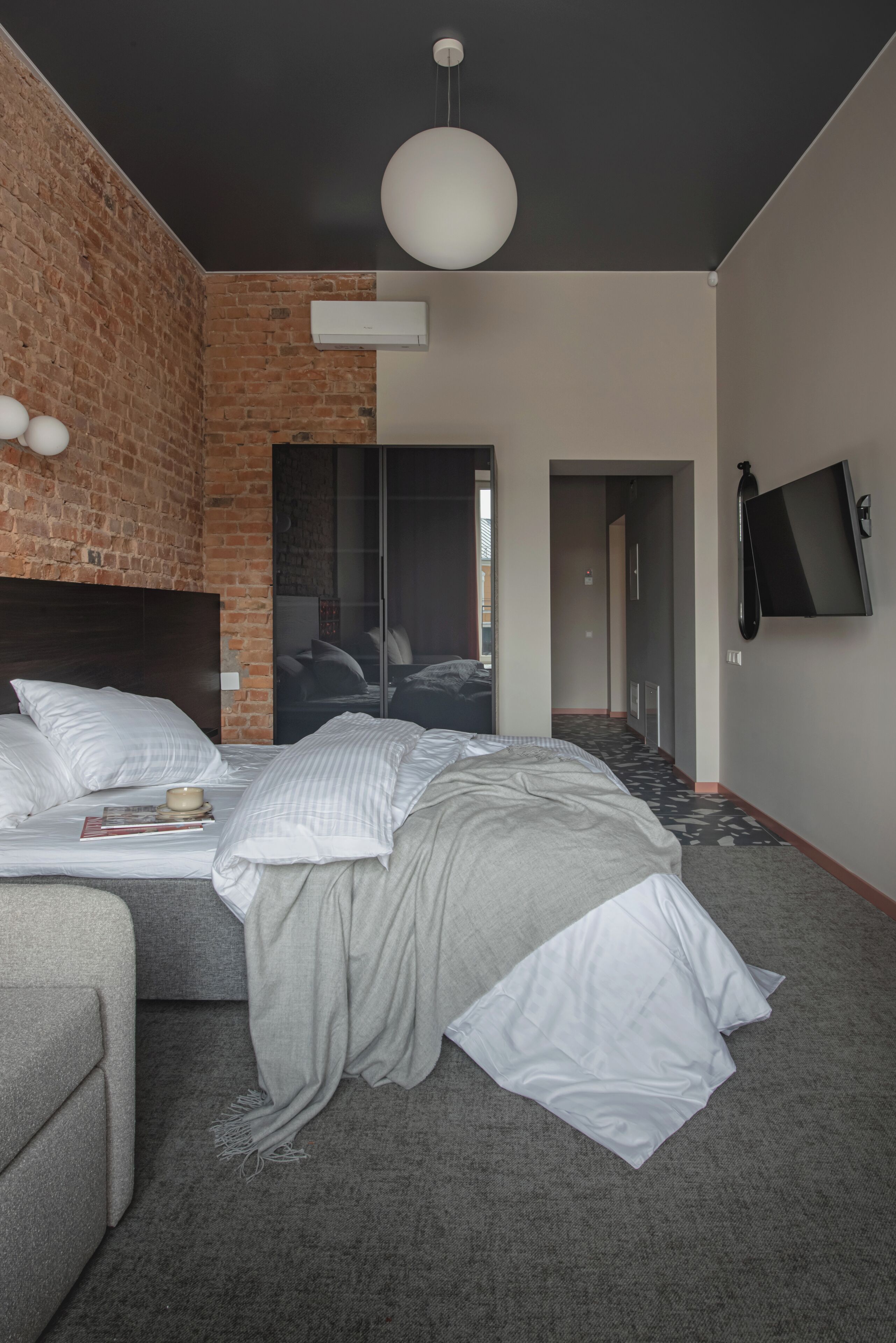 A modern hotel room with a brick accent wall, a large bed with white linens, a grey sofa, a flat-screen TV mounted on the wall, and a large pendant light.