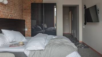 A modern hotel room with a brick accent wall, a large bed with white linens, a grey sofa, a flat-screen TV mounted on the wall, and a large pendant light.