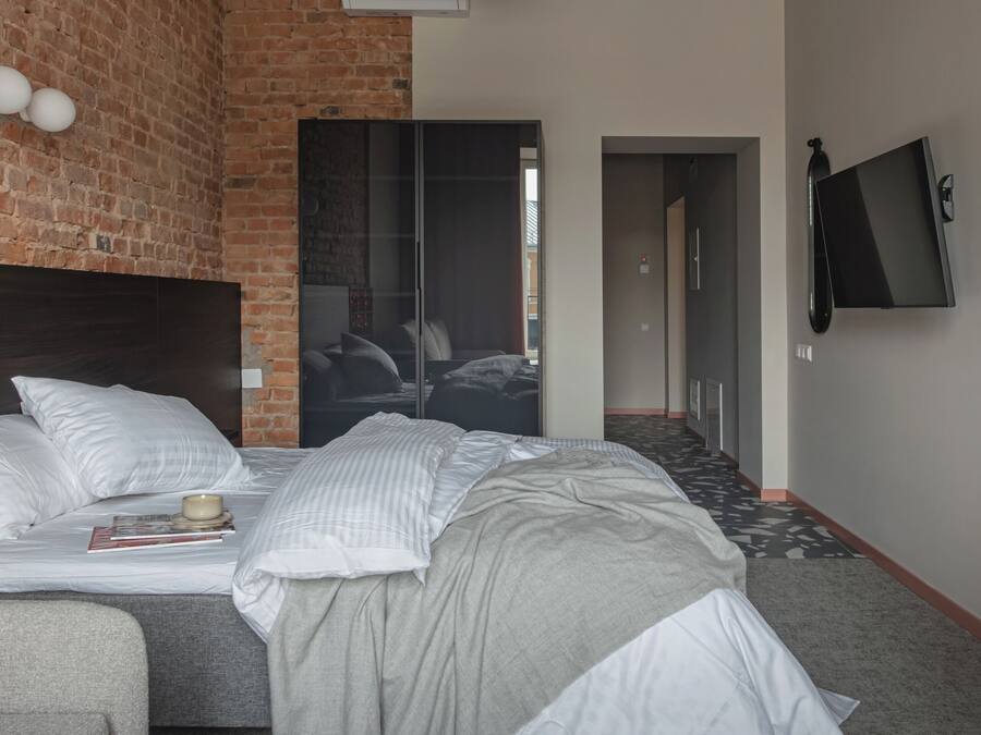 A modern hotel room with a brick accent wall, a large bed with white linens, a grey sofa, a flat-screen TV mounted on the wall, and a large pendant light.