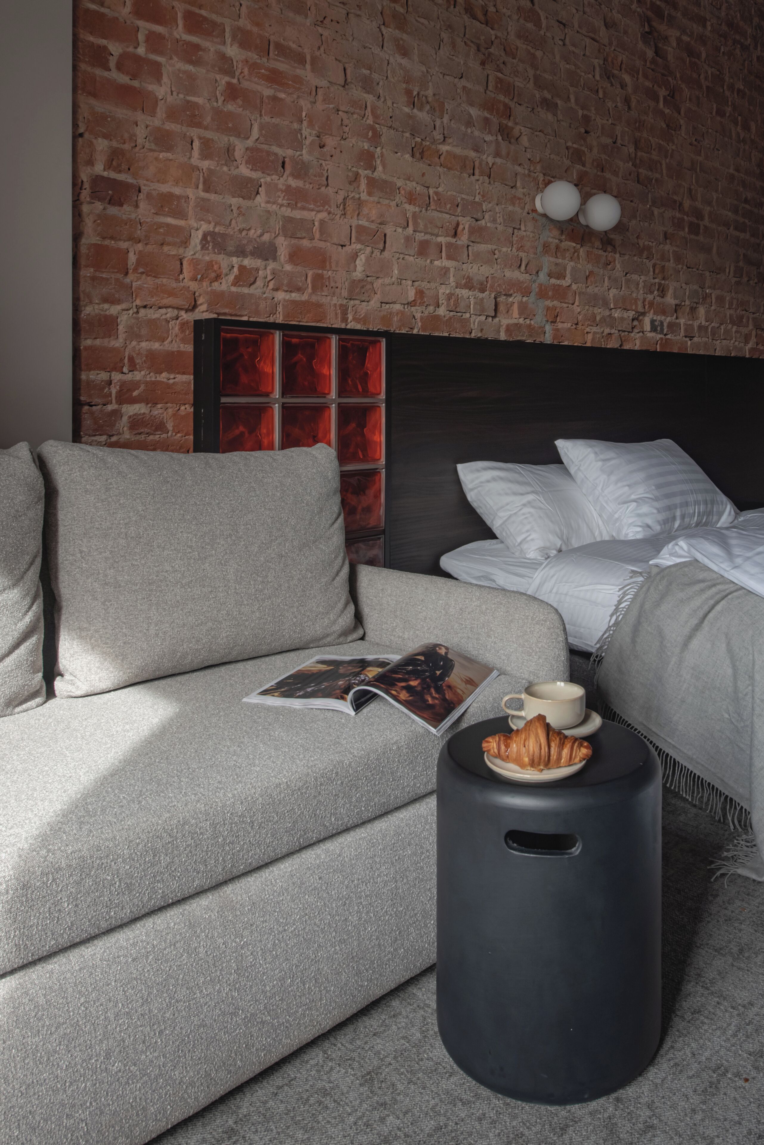 Una habitación moderna con pared de ladrillo, un sofá, una cama con ropa blanca, una revista, una taza y un croissant sobre un taburete negro.