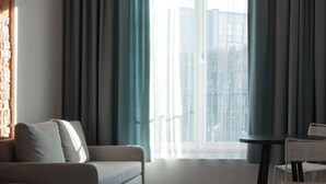 A hotel room with a bed, a sofa, a small table, and a window with curtains. - Resume Apartments (Kaunas)