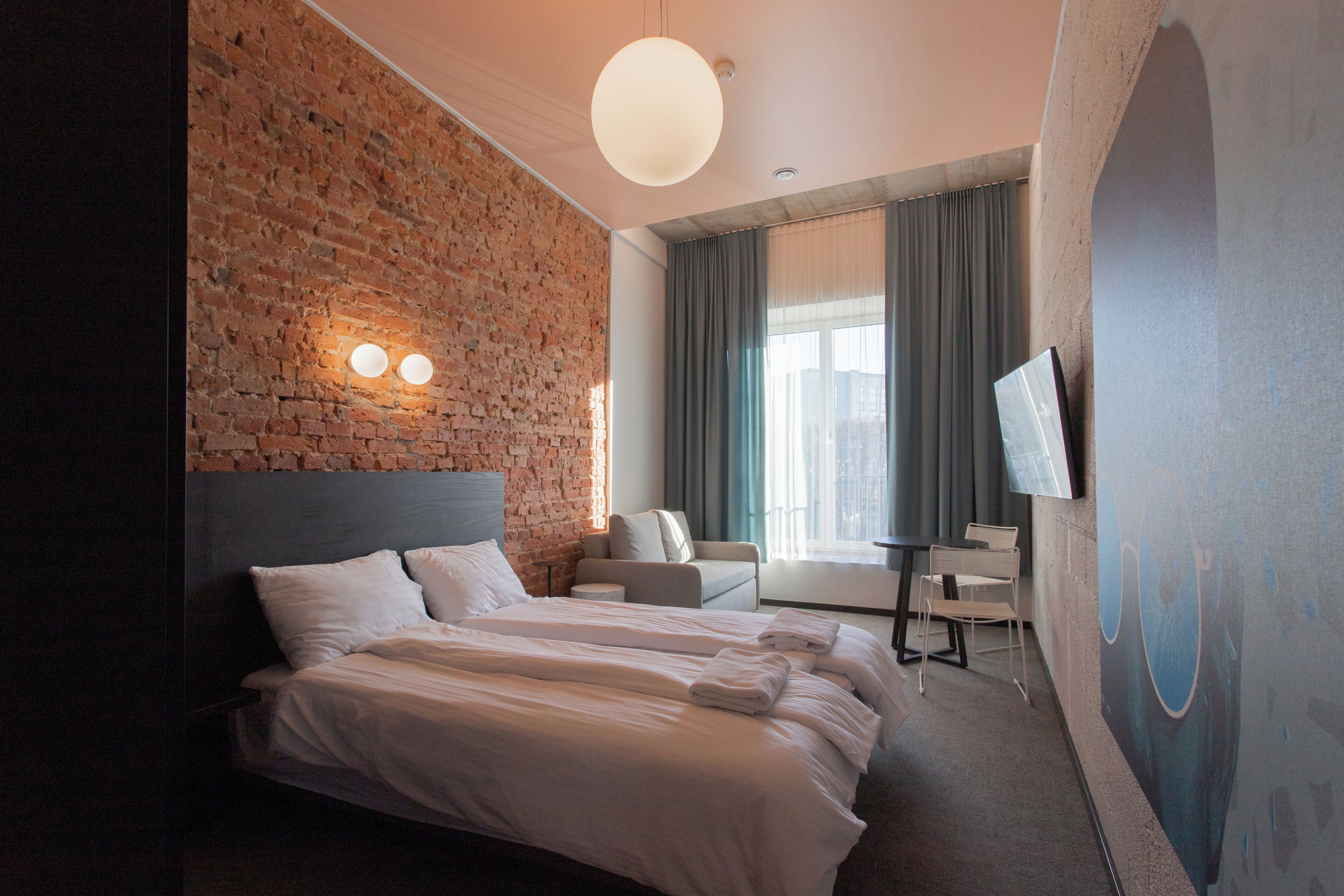 Ett hotellrum med en tegelvägg, en stor säng, en soffa, en tv och ett fönster med gardiner.
