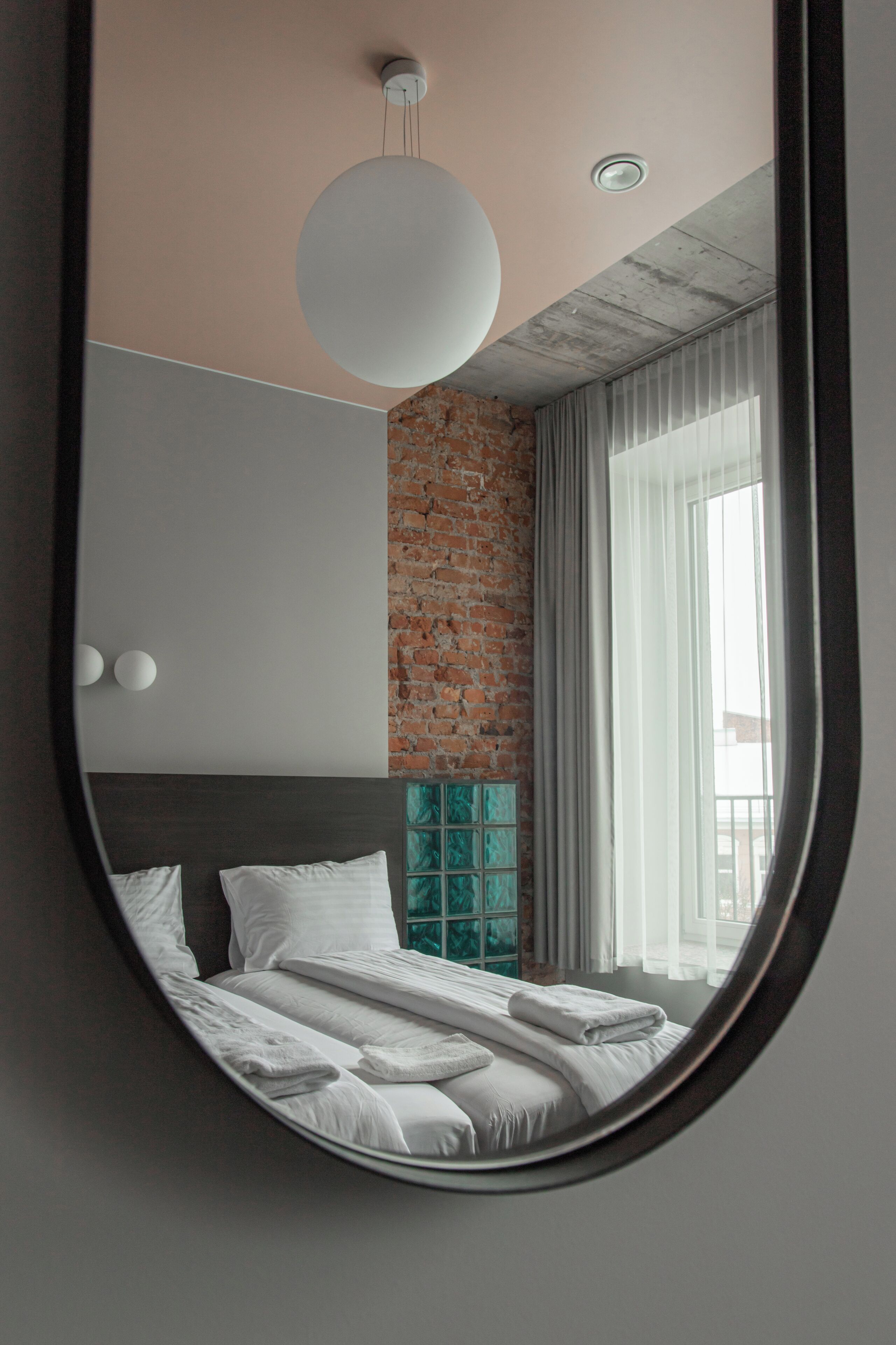 Un dormitorio con pared de ladrillo, un espejo redondo grande, cama con ropa blanca y ventana con cortinas.