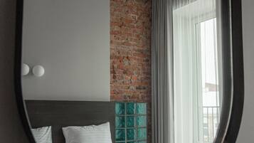Un dormitorio con pared de ladrillo, un espejo redondo grande, cama con ropa blanca y ventana con cortinas.