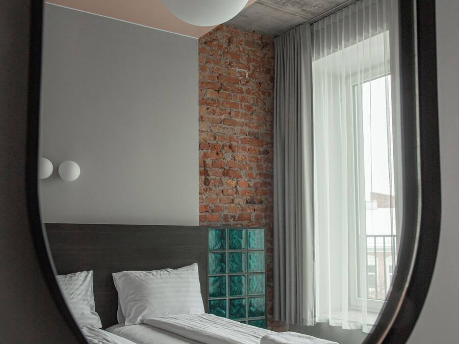 Un dormitorio con pared de ladrillo, un espejo redondo grande, cama con ropa blanca y ventana con cortinas.