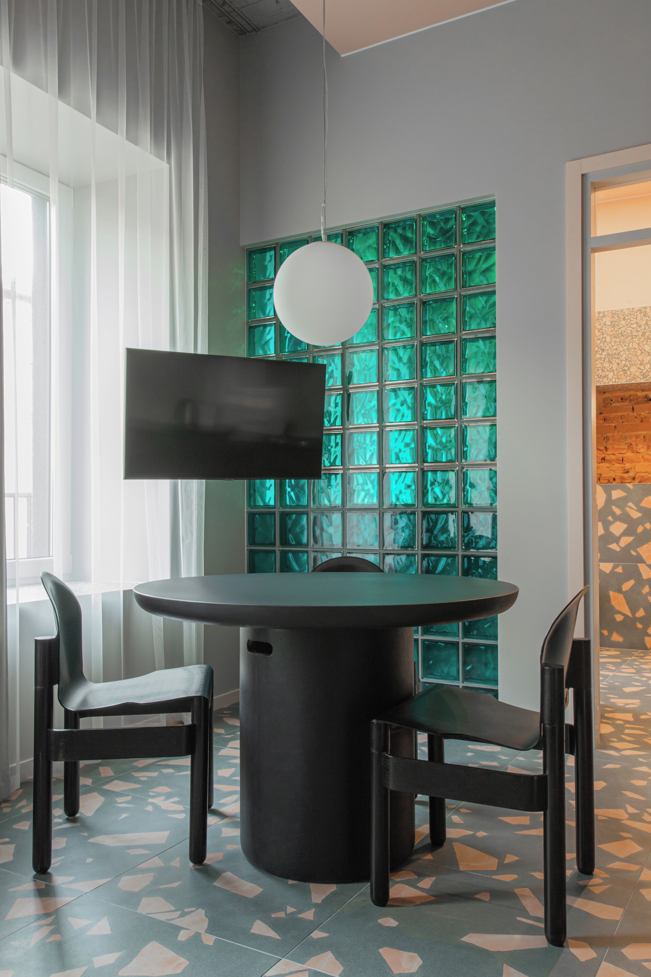 A modern dining area with a round table, two chairs, a flat-screen TV, and a green-tinted glass partition.