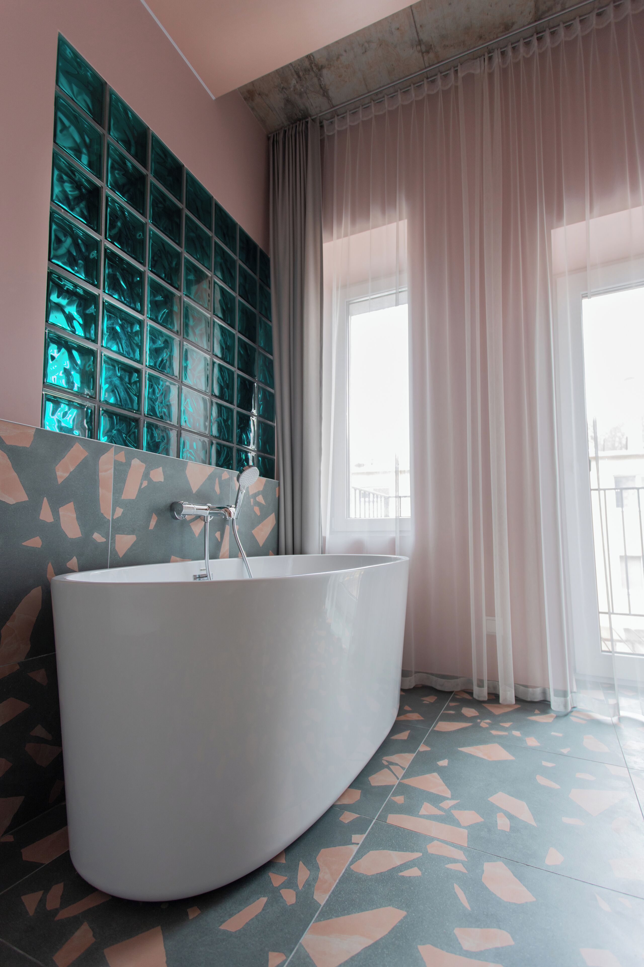 A modern bathroom with a freestanding bathtub, a large window with sheer curtains, and a terrazzo floor.