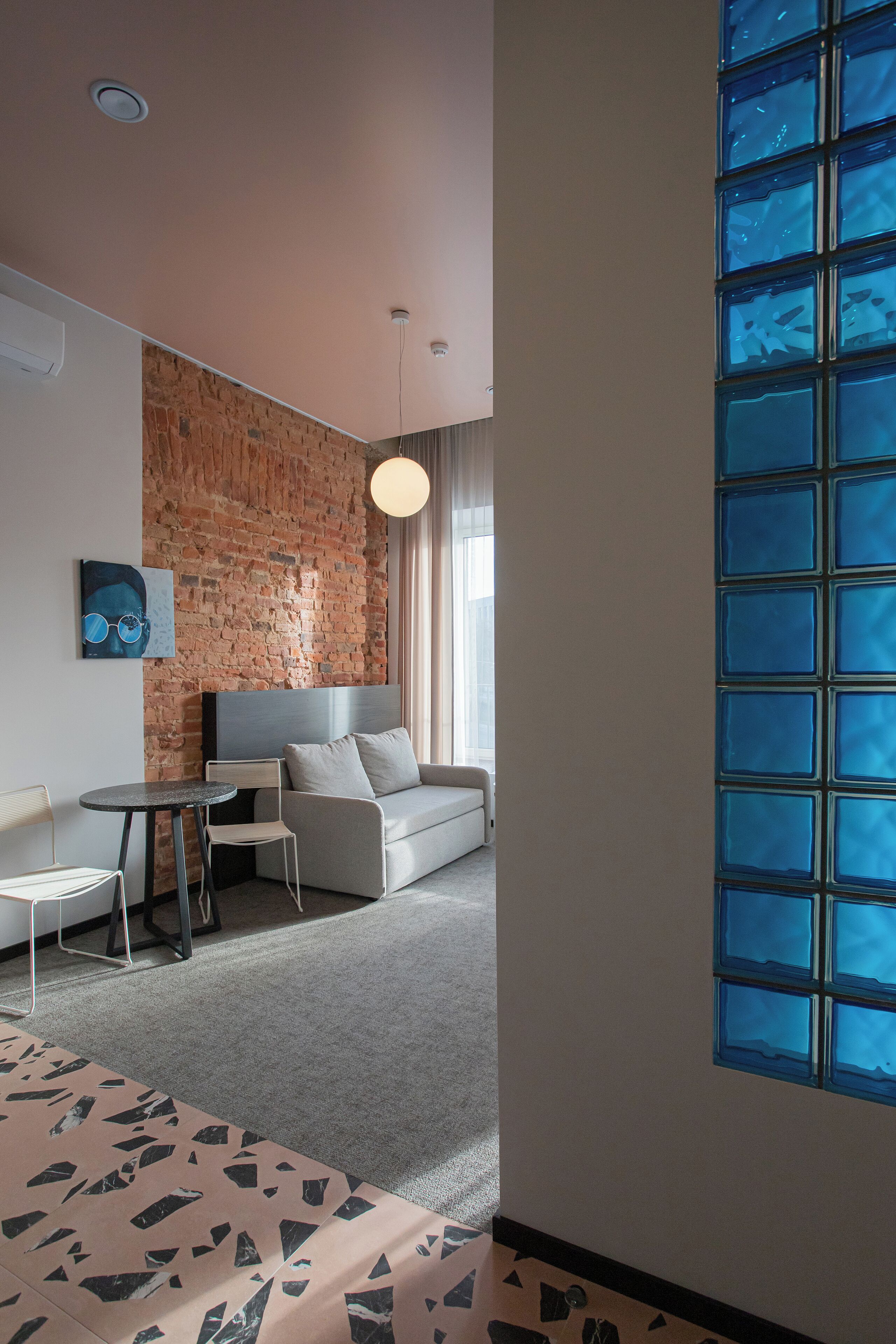 A modern hotel room with a brick wall, a grey sofa, a small table, and a large window with blue-tinted glass.