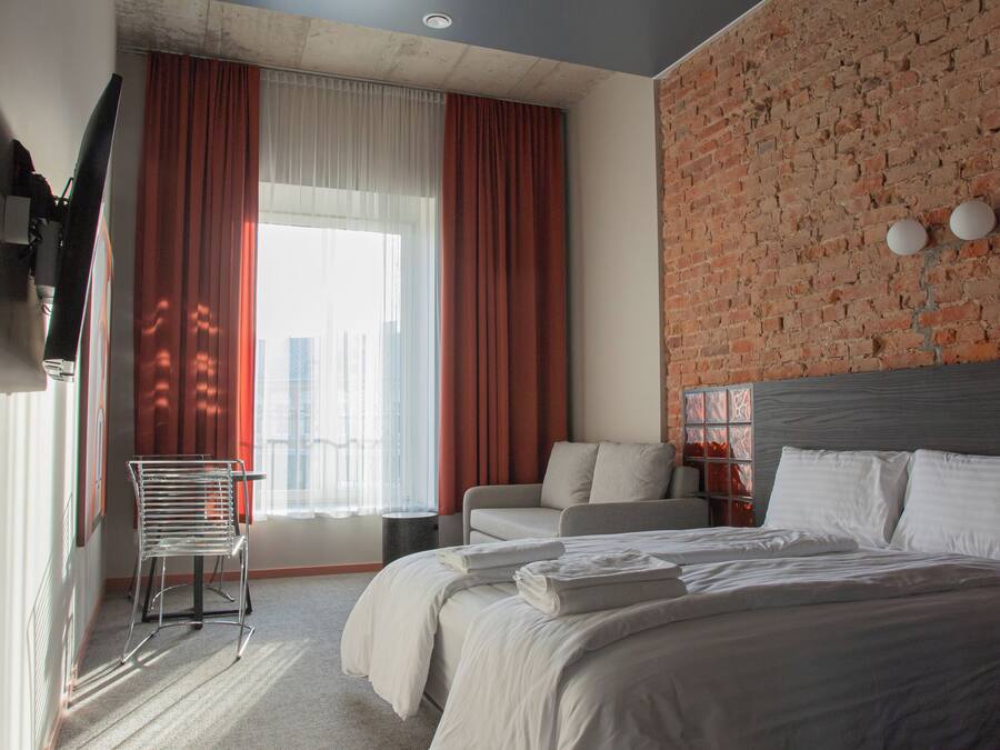 Une chambre d’hôtel moderne dotée d’un grand lit, d’un coin salon et offrant une vue sur la ville grâce à de grandes fenêtres.