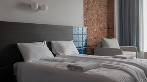A modern hotel room with a large bed, a seating area, a brick accent wall, and a large window with curtains. - Resume Apartments (Kaunas)