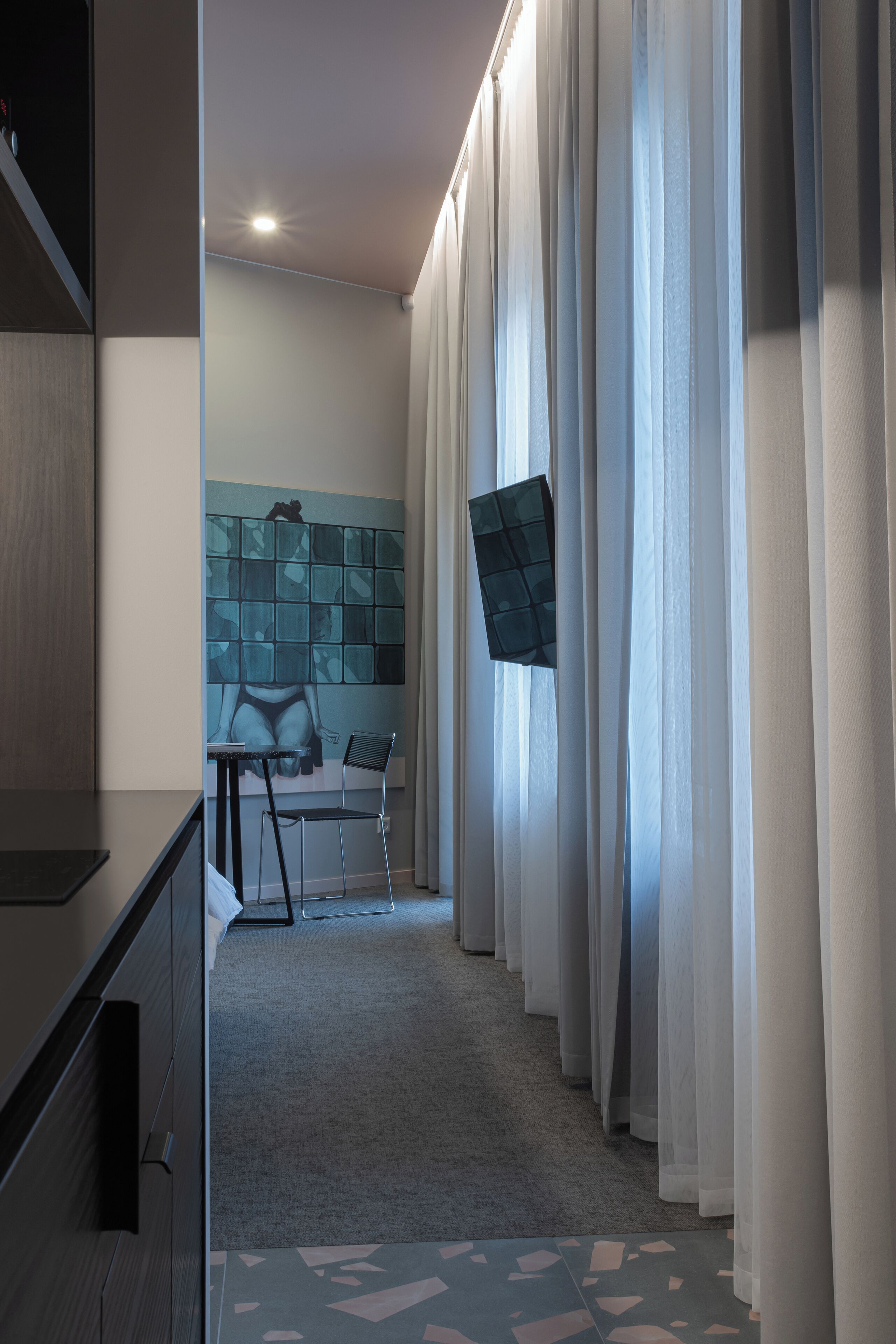 A hotel room with a bed, a desk, and a chair. There is a window with curtains, a wall with a geometric pattern, and a carpeted floor.