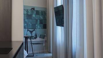 A hotel room with a bed, a desk, and a chair. There is a window with curtains, a wall with a geometric pattern, and a carpeted floor.