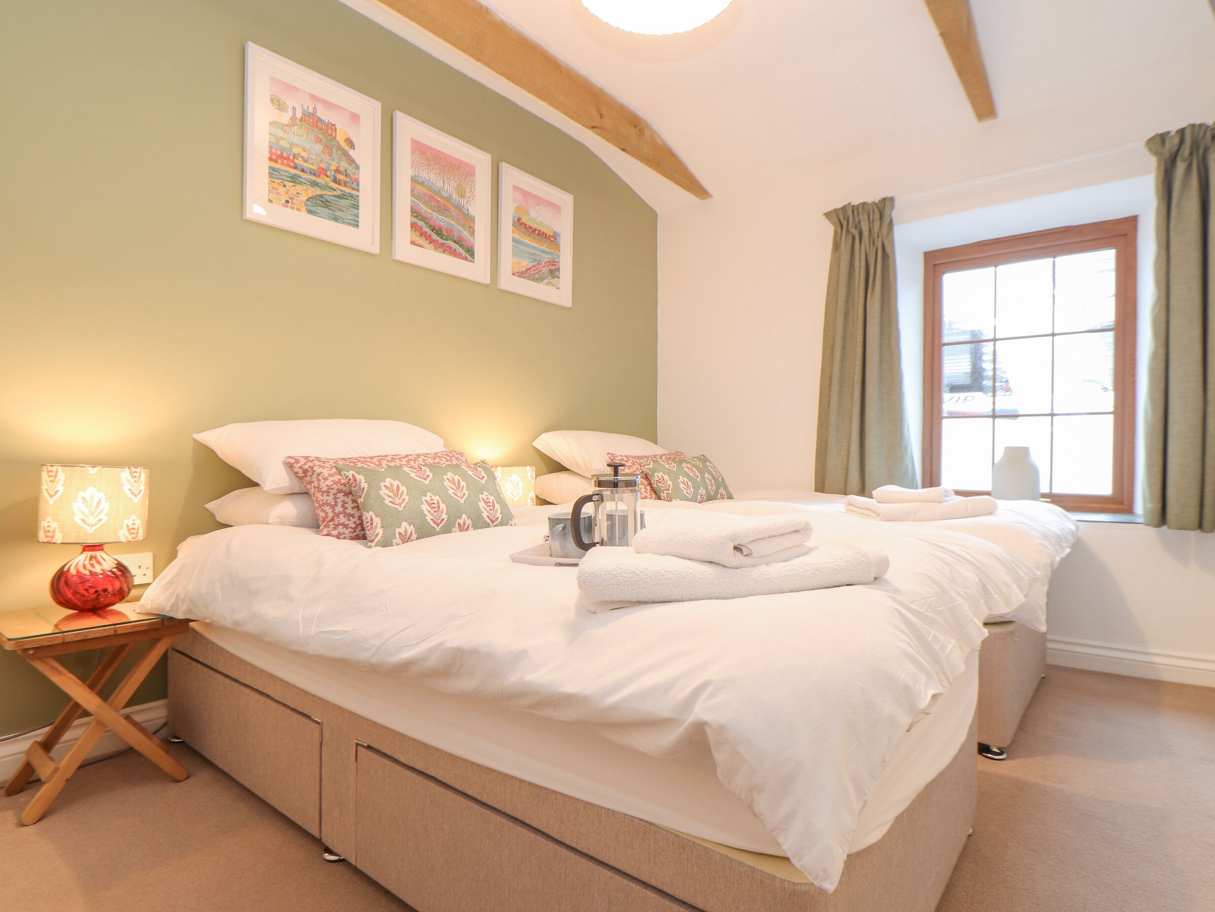 A well-lit bedroom with a large bed, a bedside table with a lamp, and a window with curtains.