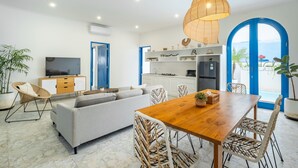 Un salon moderne comprenant un canapé, une table à manger et des chaises, un coin cuisine avec des meubles blancs et un grand téléviseur.