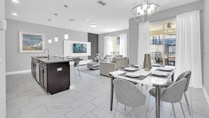 Eine moderne Küche mit Kochinsel, ein Essbereich mit Esstisch für vier Personen und ein Wohnzimmer mit Sofa und Fernseher.