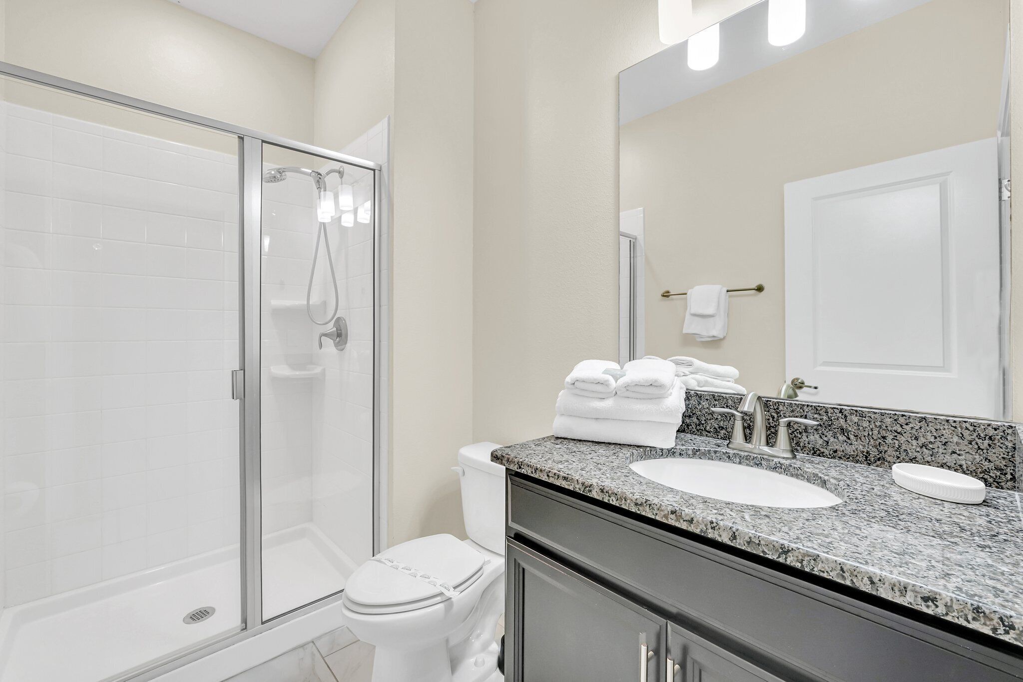 Une salle de bain moderne avec une douche à parois de verre, des toilettes blanches, un lavabo sur un plan de travail en granit et un grand miroir.