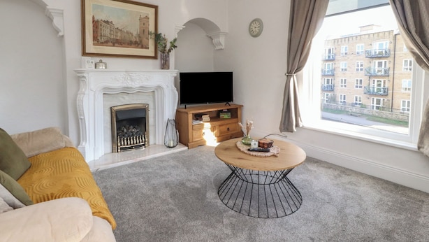 A living room with a fireplace, a television, a round coffee table, and a large window with curtains. - Bridge End (Skipton)