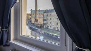 A view through a window with curtains, featuring a canal with boats and a tall chimney of a building in the background. - Bridge End (Skipton)
