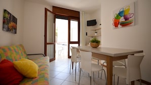 A modern living room with a colorful sofa, a dining table with chairs, a TV mounted on the wall, and a door leading to an outdoor area. - Nicely furnished flat in the city centre (Bibione Pineda)