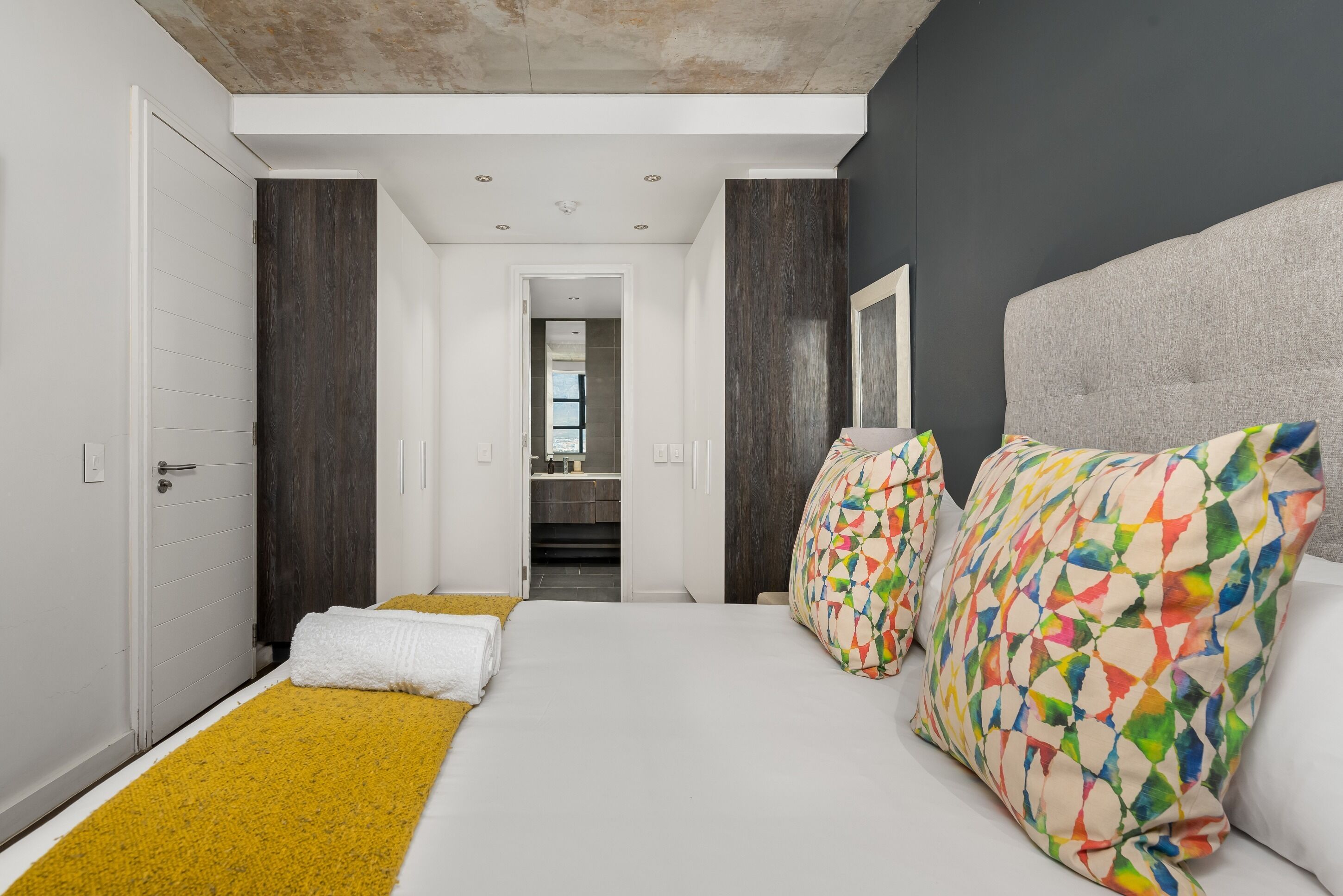 A modern hotel room with a large bed, a yellow throw rug, and colorful pillows.