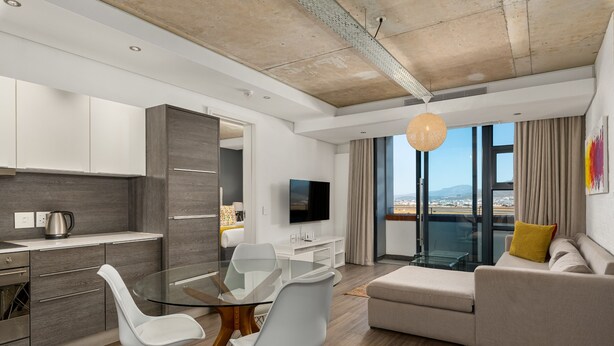 A modern living room with a glass dining table, white chairs, a beige sofa, and a TV on the wall. - Modern 1 Bedroom Apartment in Century City (Century City)