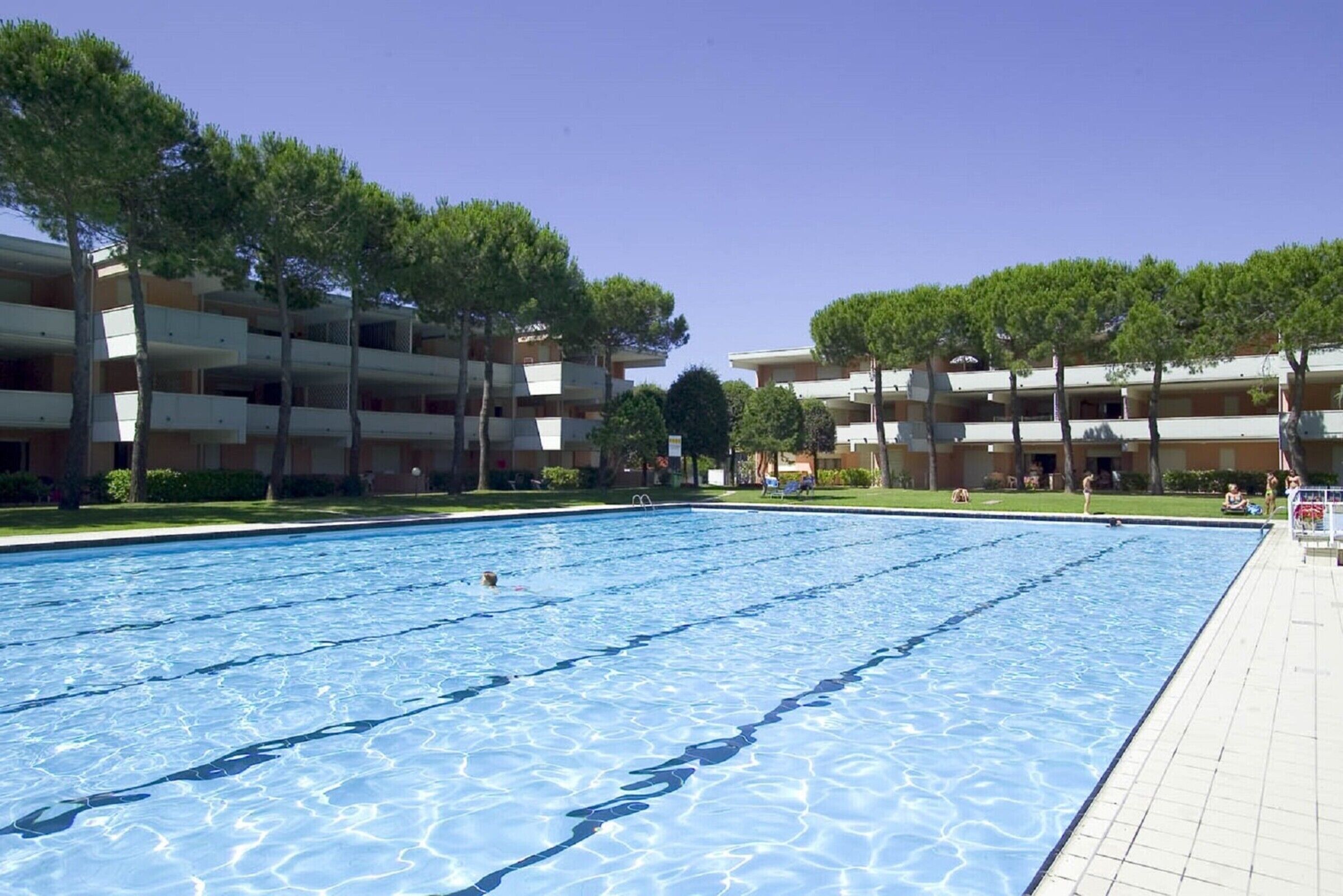 A swimming pool surrounded by multi-story buildings with balconies, trees, and a clear sky.