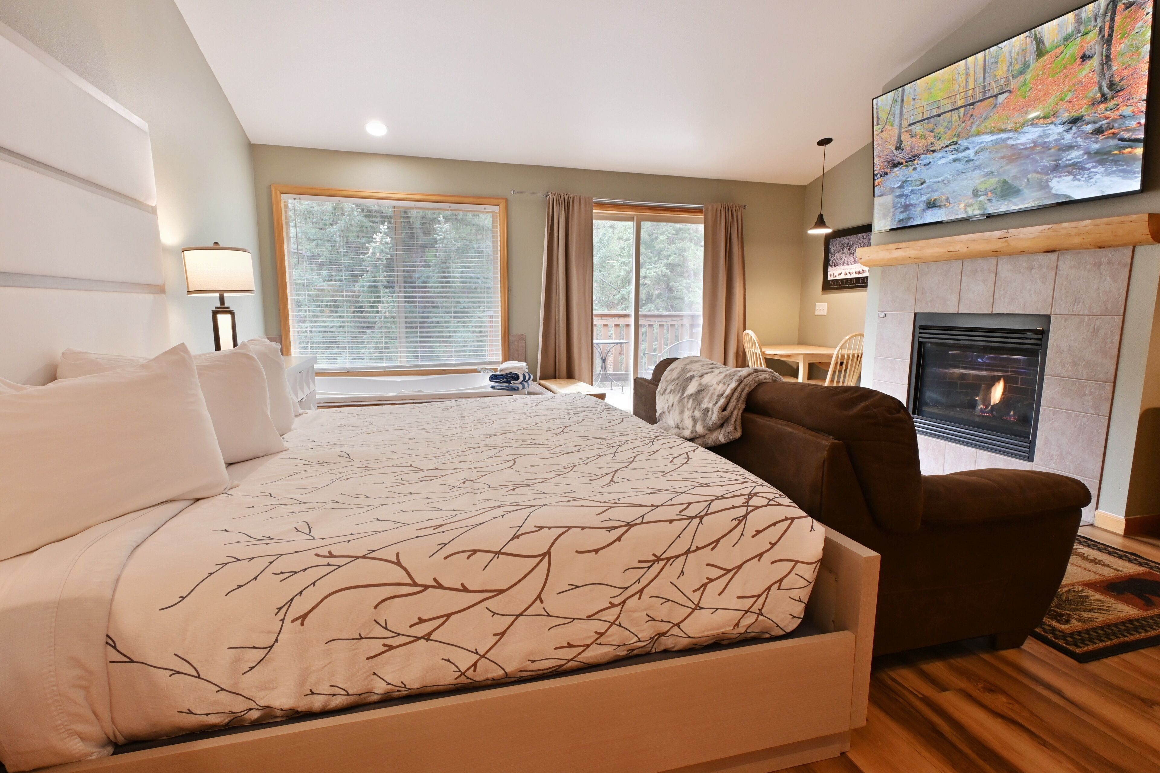 A bedroom with a large bed, a fireplace, a TV mounted on the wall, and a balcony with a view of trees and a bridge.
