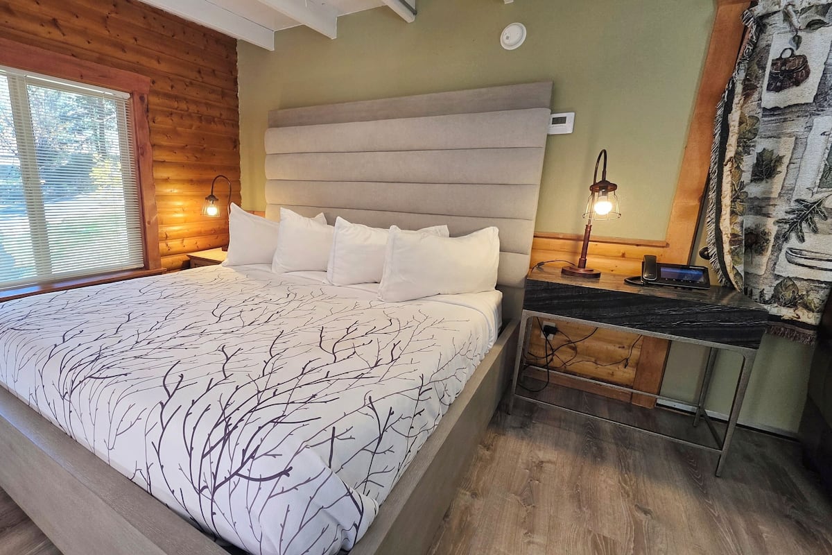 Une chambre à coucher de style cabane en bois, avec un grand lit, une table de chevet avec une lampe et une fenêtre équipée de stores.