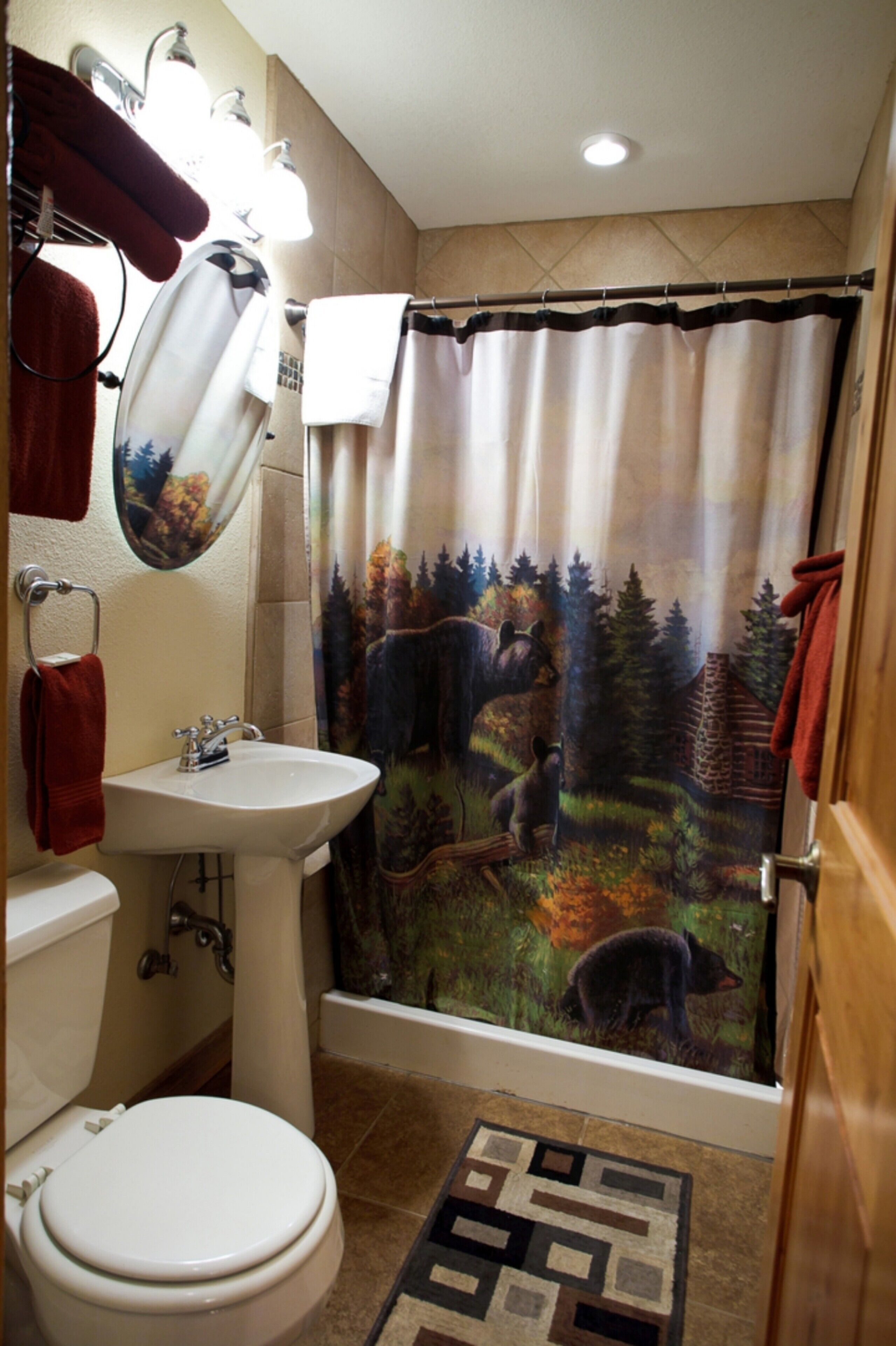A bathroom with a white sink, a toilet, a shower curtain with a forest scene, and a patterned rug on the floor.