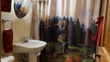 Un baño con un lavamanos blanco, un inodoro, una cortina de ducha con un paisaje boscoso y una alfombra estampada en el suelo.