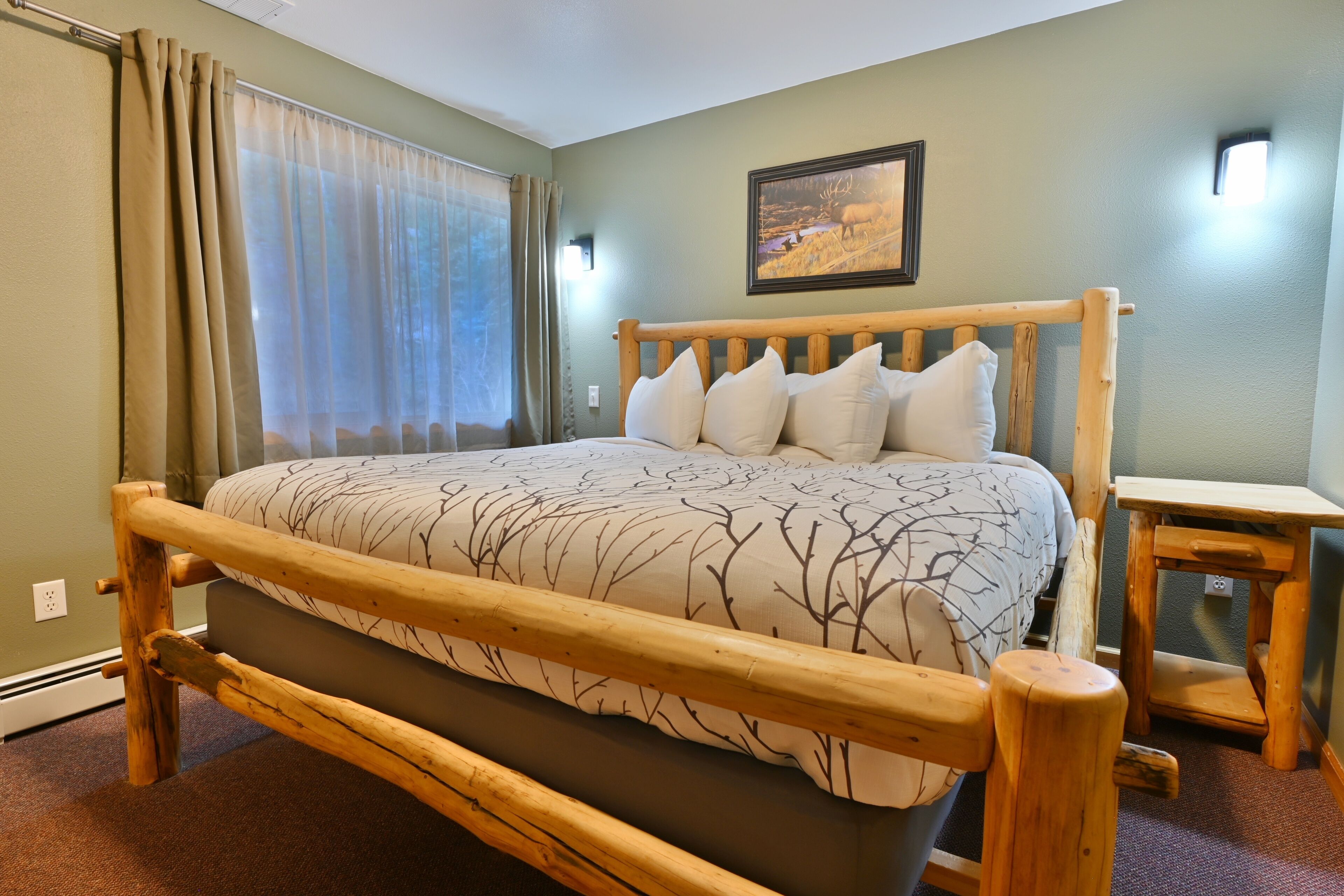 A wooden bed with a tree branch pattern bedspread, a wooden nightstand, and a painting on the wall.