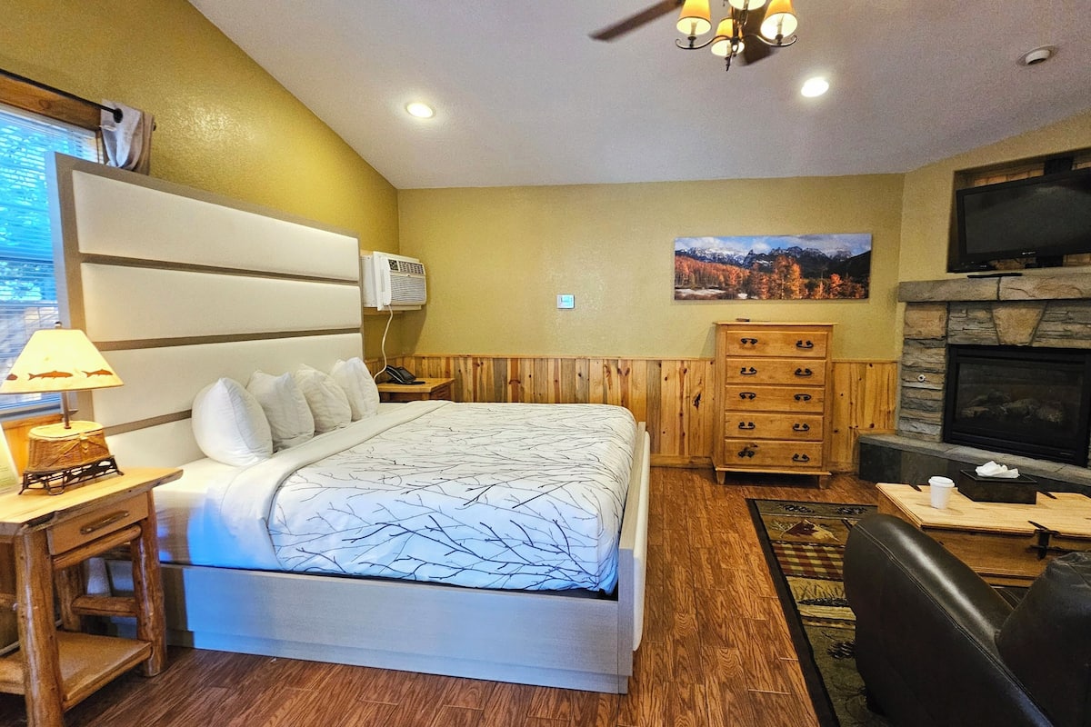 Une chambre d’hôtel avec un grand lit, une commode en bois, une télévision, une cheminée et un bureau avec une lampe.
