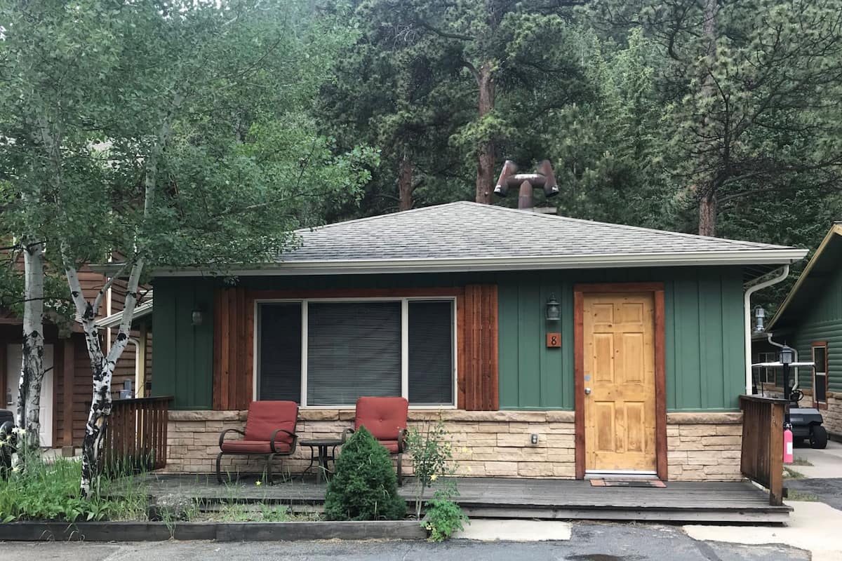 Une maison de plain-pied à l’extérieur vert, avec une porte en bois et deux chaises rouges devant.