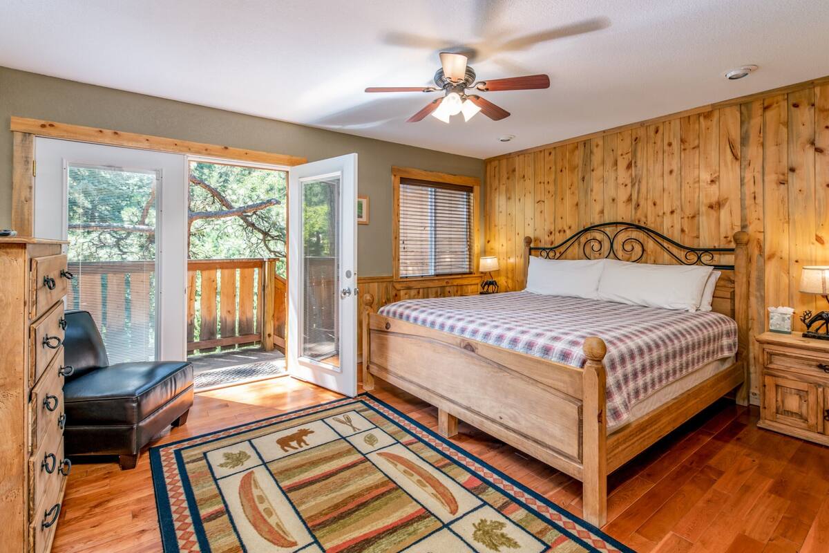 Une chambre avec un lit en bois, un ventilateur de plafond, une table de chevet, une commode et une vue sur l’extérieur grâce à une porte-fenêtre coulissante.
