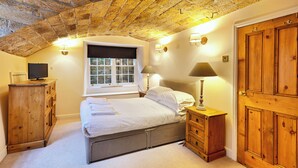 A bedroom with a stone ceiling, a bed, a wooden dresser, a nightstand with a lamp, a window with a blind, and a wooden wardrobe. - The Old Wine Cellar - 4 bedroom property (Oakerthorpe)