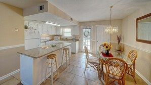 A kitchen with white cabinets, a granite countertop, and a dining area with wicker chairs and a vase of sunflowers. - 206 White Sandy-pet Friendly (Panama City Beach)